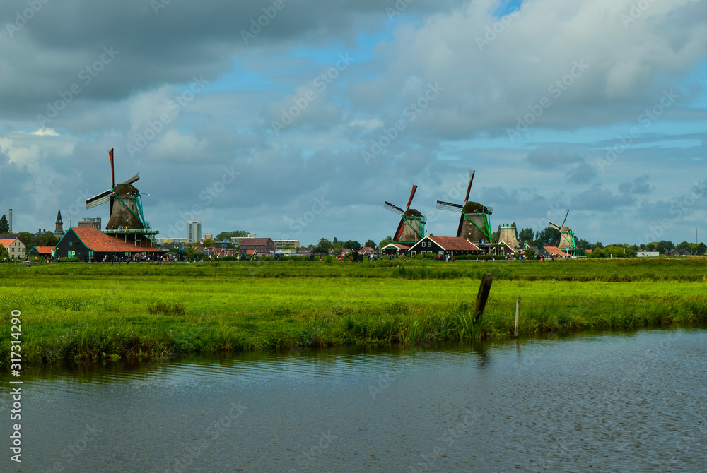 Fototapeta premium Zaanse Schans, Holland, August 2019. North-east of Amsterdam is a small community located on the quay of the Zaan river. View from above of the fields with mills, tourists are noticed. Cloudy day.