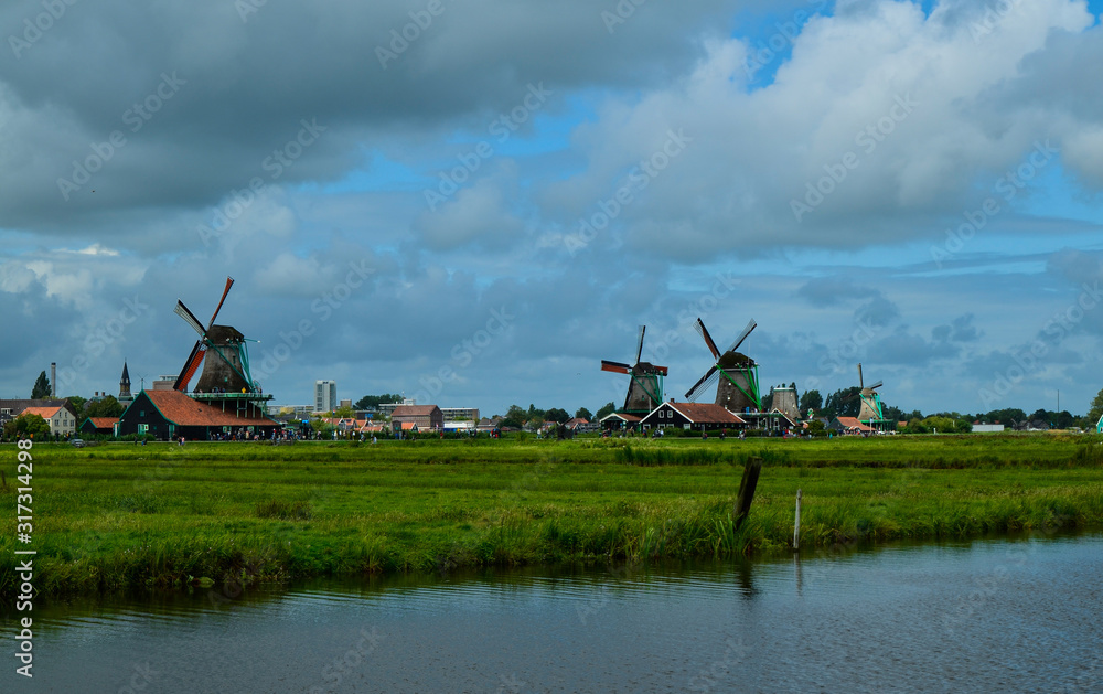 Fototapeta premium Zaanse Schans, Holland, August 2019. North-east of Amsterdam is a small community located on the quay of the Zaan river. View from above of the fields with mills, tourists are noticed. Cloudy day.
