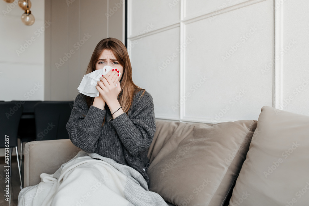 ill woman blowing nose with tissue sitting on couch at home in apartment - obrazy, fototapety, plakaty 