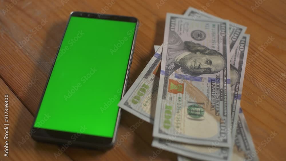 Man folds hundred dollar bills next to phone greenscreen touchpad on table. Mock-up with green screen cellphone. Money cash chroma key concept. Technology and economy. Personal budget. Home finance. Man folds hundred dollar bills next to phone greenscreen touchpad on table. Mock-up with green screen cellphone. Money cash chroma key concept. Technology and economy. Personal budget. Home finance.