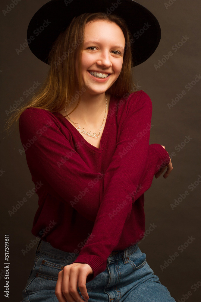 Studio portrait of young blonde girl in a black hat, red blouse and blue jeans on a brown background. Studio portrait of young blonde girl in a black hat, red blouse and blue jeans on a brown background.