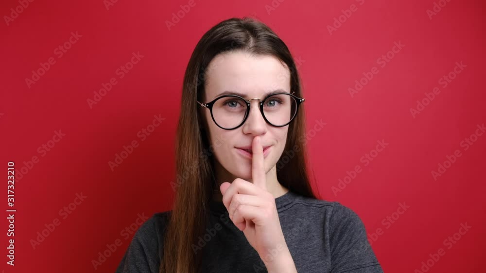 Tender beautiful young female with pleased expression makes shush gesture, has gentle smile, gossip wears glasses and grey t-shirt, stands over red background. Tender beautiful young female with pleased expression makes shush gesture, has gentle smile, gossip wears glasses and grey t-shirt, stands over red background.