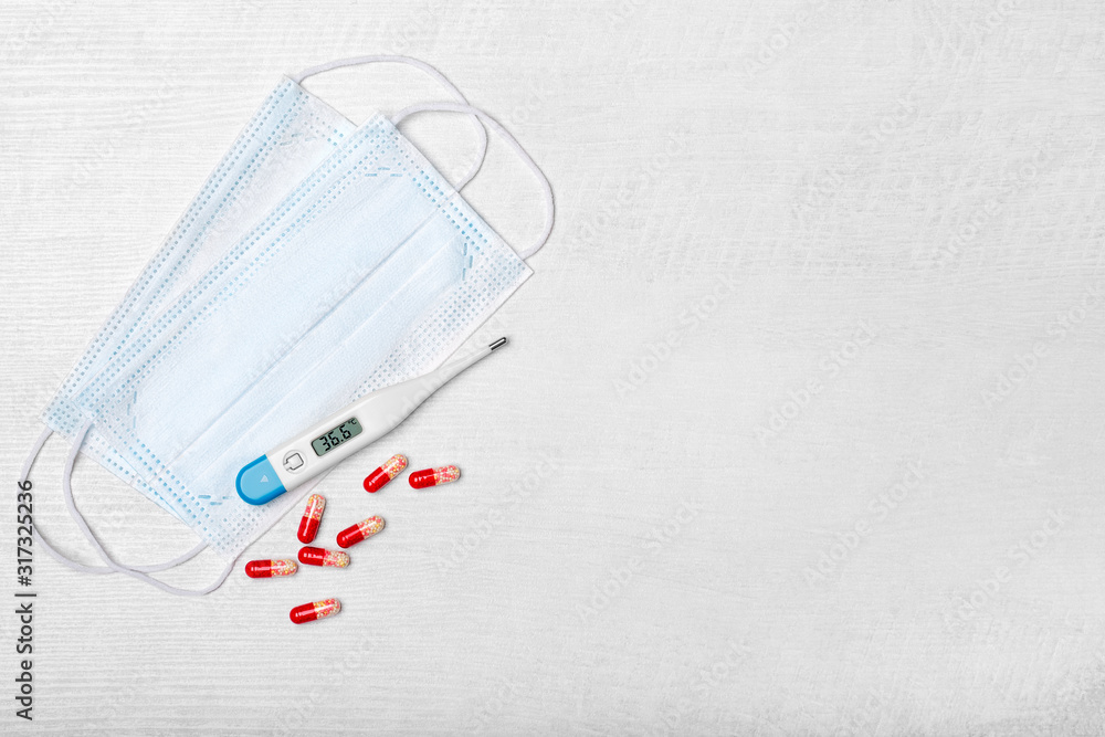 Creative flat lay overhead composition with disposable face masks, electronic body thermometer, and scattered pills on a light wooden background. Top view. Free copy space for text Creative flat lay overhead composition with disposable face masks, electronic body thermometer, and scattered pills on a light wooden background. Top view. Free copy space for text