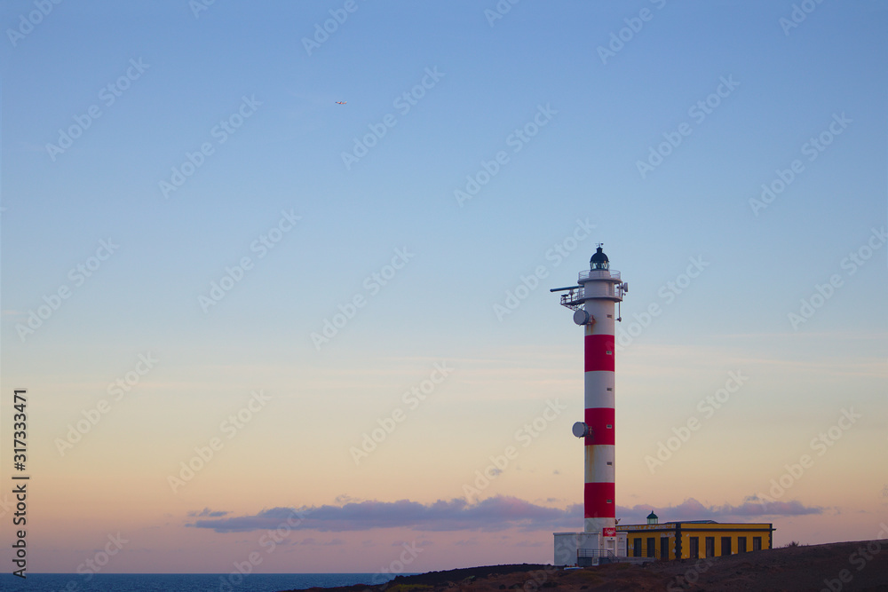 A lighthouse of white and red colors stands out on the reddish horizon of a sunset