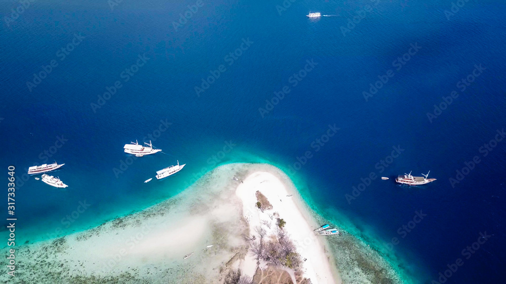 Top down drone shot of a paradise island with some boats anchored around in Komodo National Park, Flores, Indonesia. Brownish island turns into white sand beach and further into turquoise and navy sea