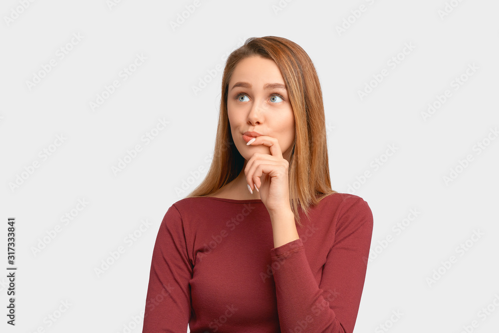 Girl looks away, touches chin and twists lips. Beautiful young woman wears Burgundy-colored jacket sleeves isolated on white background in Studio with an empty space for text and advertising. Girl looks away, touches chin and twists lips. Beautiful young woman wears Burgundy-colored jacket sleeves isolated on white background in Studio with an empty space for text and advertising.