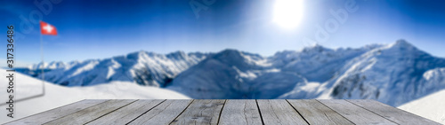 wooden shelf display table top against blue winter mountain panorama and swiss flag snow covered blue mountain layers © Simon