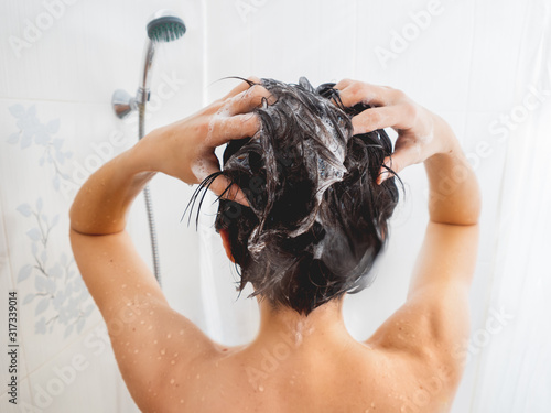 Naked woman with short hair takes a shower. Woman washes her hair with shampoo. White bathroom.