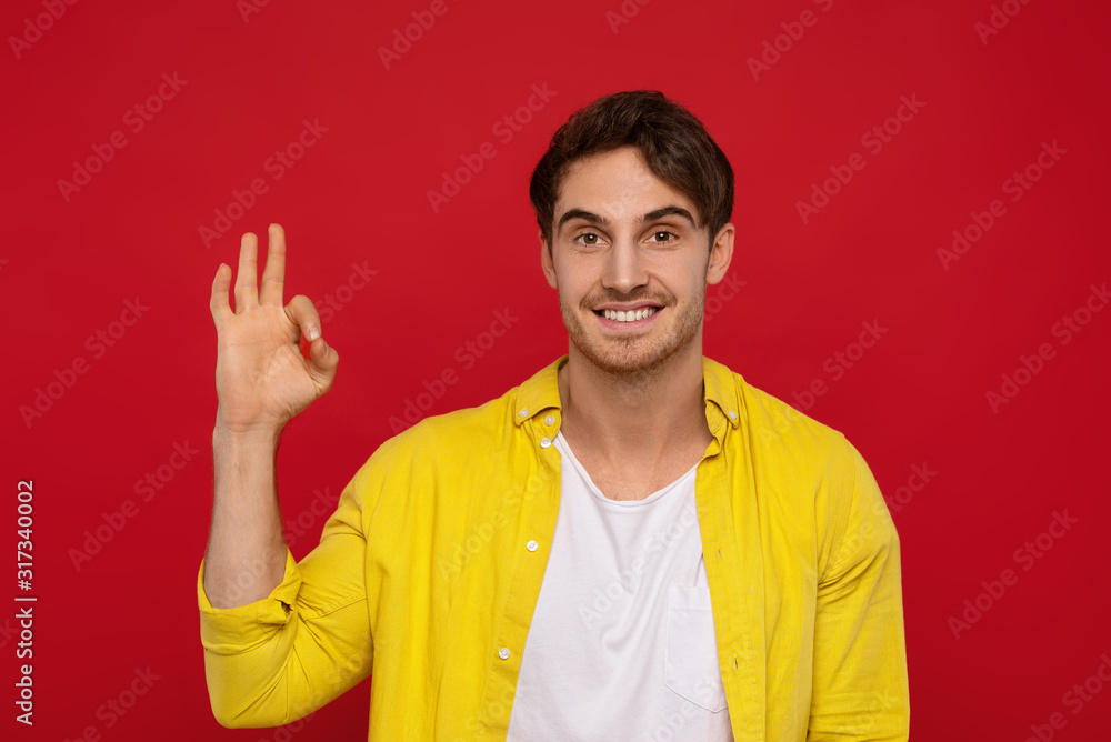Yeah, it's ok. Positive handsome man in yellow shirt showing ok sign with one hand, has big smile, being in high spirit, isolated on red background Yeah, it's ok. Positive handsome man in yellow shirt showing ok sign with one hand, has big smile, being in high spirit, isolated on red background