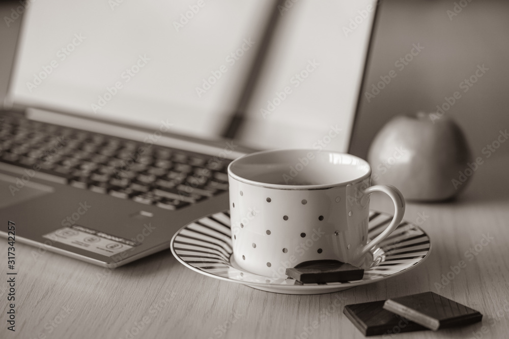 Cup of coffee on the wooden table with notebook on the background. Piece of black chocolate is on the saucer, break time in the office for refreshment Cup of coffee on the wooden table with notebook on the background. Piece of black chocolate is on the saucer, break time in the office for refreshment