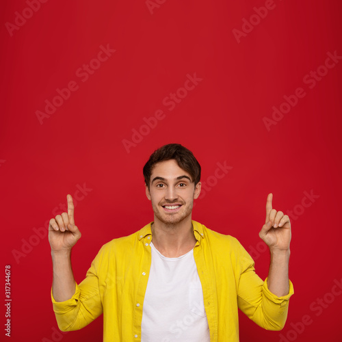 Cheerful handsome man in yellow shirt with amazed happy expression pointing with fingers up isolated on red background. Man in pleased to notice something © MarHo