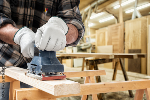 Close-up. Carpenter with his hands protected by gloves with the electric sander smoothes a wooden board. Construction industry, carpentry workshop.