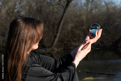 Woman with dark long hair in black robes is holding polished Clear Quartz Sphere in her hands in front of the lake.