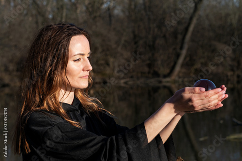 Woman with dark long hair in black robes is holding polished Shungite Sphere in her hands in front of the lake.