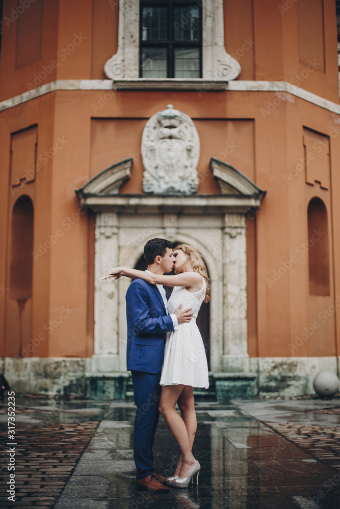 Stylish couple  kissing in european city street on background of old architecture. Fashionable bride and groom in love enjoying day, embracing in city. Traveling together in Europe