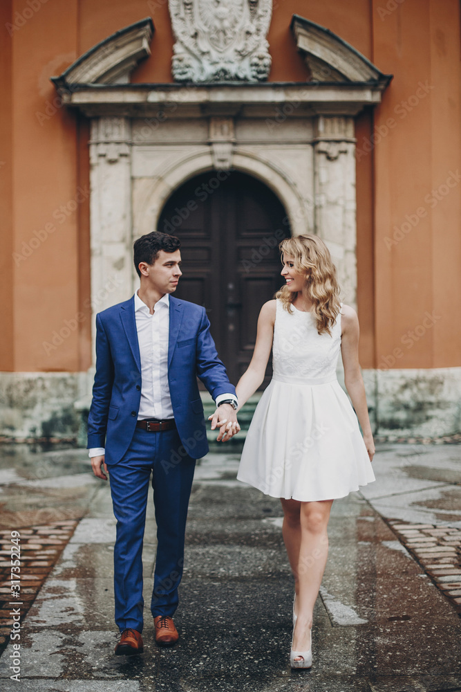 Stylish couple walking together in european city street on background of old architecture. Fashionable bride and groom in love enjoying day in city. Traveling together in Europe