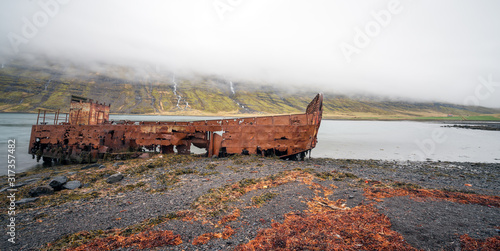 Rusty and abandoned old ship wreck from world war two laying on land in Mjoifjordur in Iceland. Waterfalls and foggy landscape scenery. Transport and ship concept.