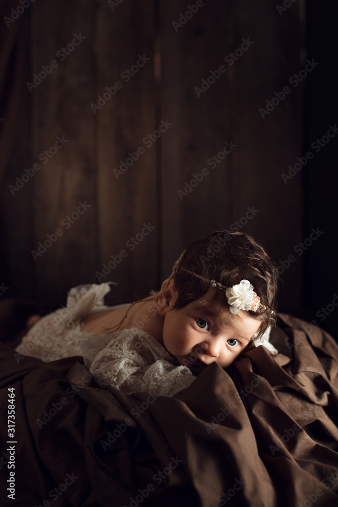 Beautiful infant in white dress, with flower headband lies on brown drapery, vintage blanket on wood background. Two month old girl. Surprised baby. Copy space