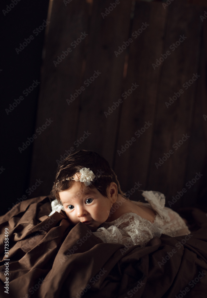 Beautiful infant in white dress, with flower headband lies on brown drapery, vintage blanket on wood background. Two month old girl. Surprised baby. Copy space