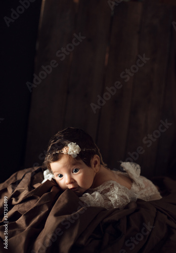 Beautiful infant in white dress, with flower headband lies on brown drapery, vintage blanket on wood background. Two month old girl. Surprised baby. Copy space
