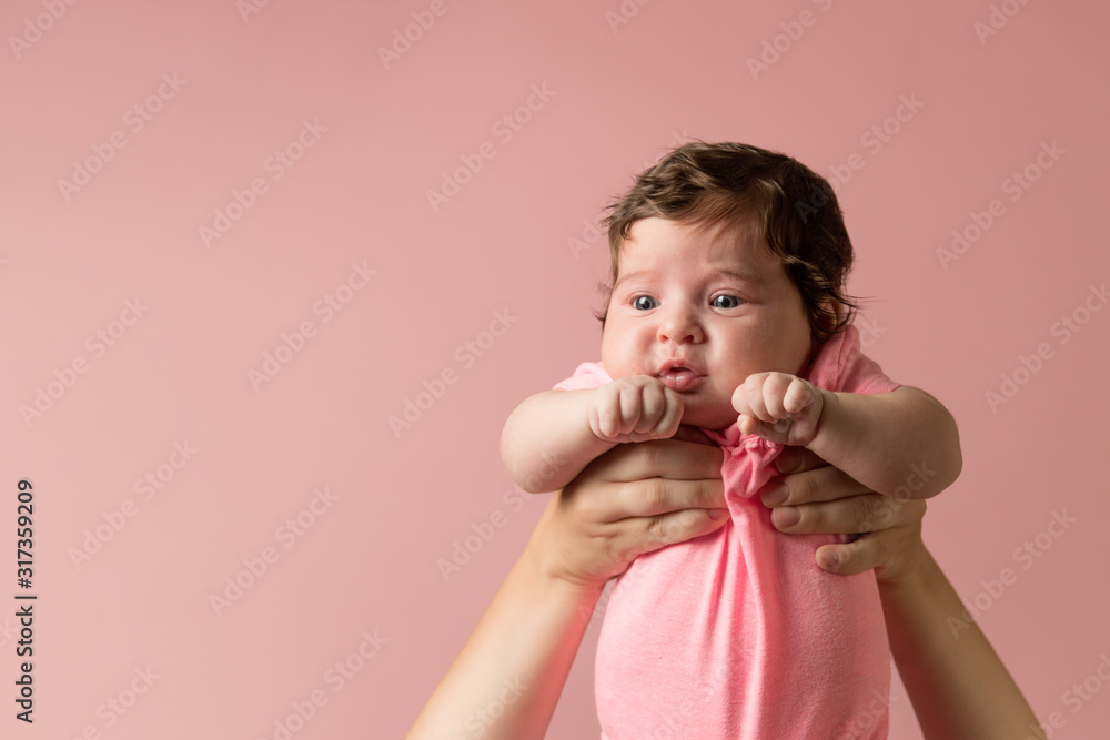 Cute infant in pink jumpsuit in mother hands on light pink background. Maternity concept. Tearfully baby. Copy space Cute infant in pink jumpsuit in mother hands on light pink background. Maternity concept. Tearfully baby. Copy space