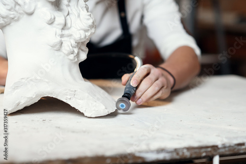 Professional bearded carver polishing with micro drill machine details of the man's sculpture head in his workshop. photo