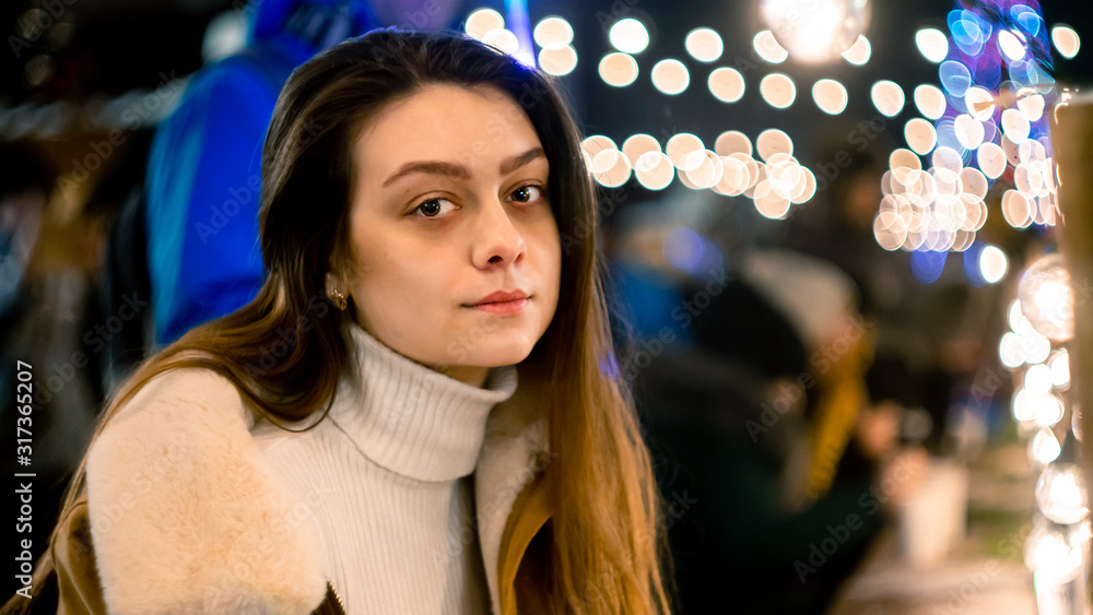 Cute girl in a Christmas park sits at a table and looks at the camera. garland background. bright lights Cute girl in a Christmas park sits at a table and looks at the camera. garland background. bright lights
