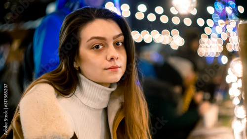 Cute girl in a Christmas park sits at a table and looks at the camera. garland background. bright lights © Дмитрий Ткачев
