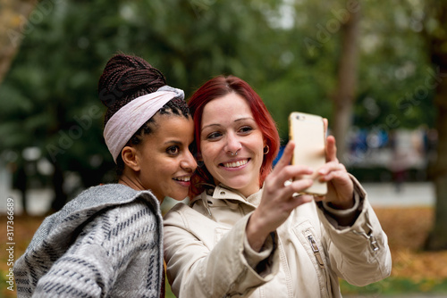 Lovely multiethnic couple of smiling women staying close and taking selfie on mobile phone in autumn park photo