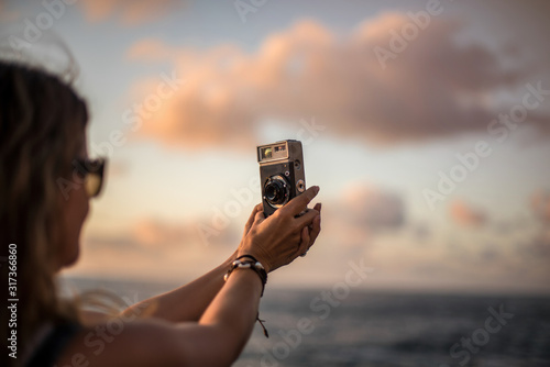 Side view of crop young woman standing at seaside at sunset time and taking picture with analog photo camera