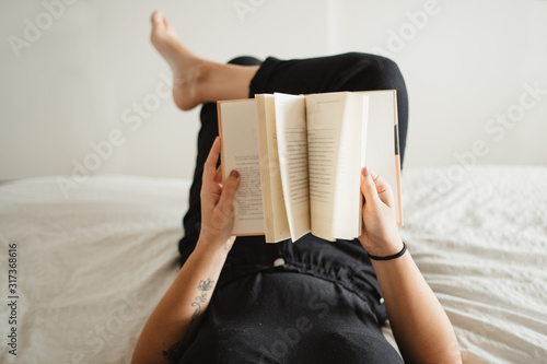Faceless female in black clothes enjoying pastime and laying on bed with legs up while reading novel at home in Paris photo
