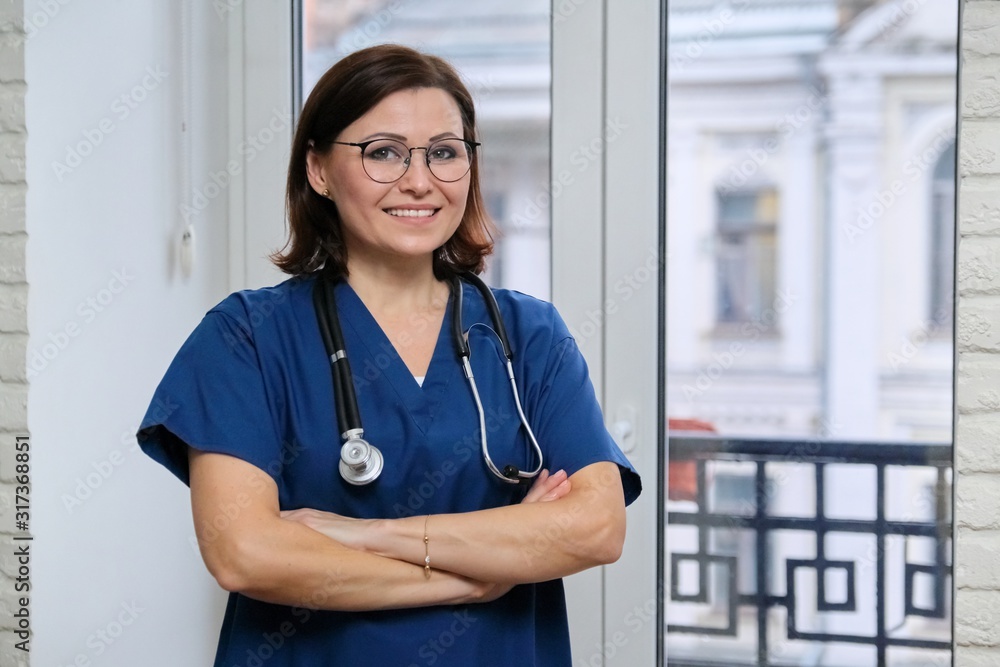 Smiling adult woman doctor in blue uniform stethoscope with folded arms, confident female medic looking at camera, standing near the window in clinic Smiling adult woman doctor in blue uniform stethoscope with folded arms, confident female medic looking at camera, standing near the window in clinic