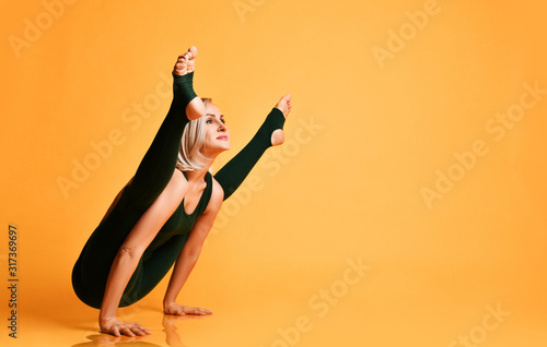 Blonde athletic girl trainer doing yoga pose asana stretches in sport fitness gym club in green sportswear on yellow 