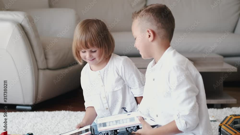Brother and sister are at home. They are playing with a toy railroad. The children are sitting on the floor. Brother and sister are at home. They are playing with a toy railroad. The children are sitting on the floor.