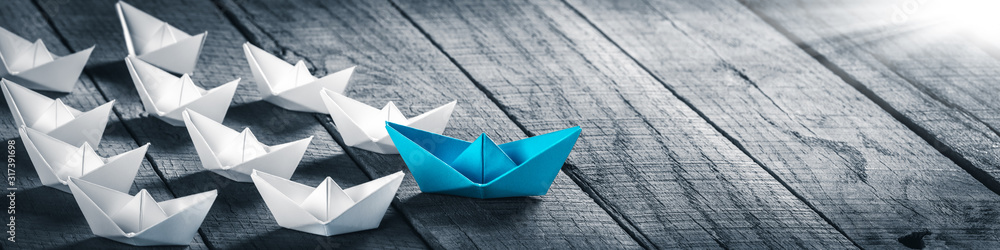 Blue Paper Boat Leading A Fleet Of Small White Boats On Wooden Table With Sunlight - Leadership Concept Blue Paper Boat Leading A Fleet Of Small White Boats On Wooden Table With Sunlight - Leadership Concept