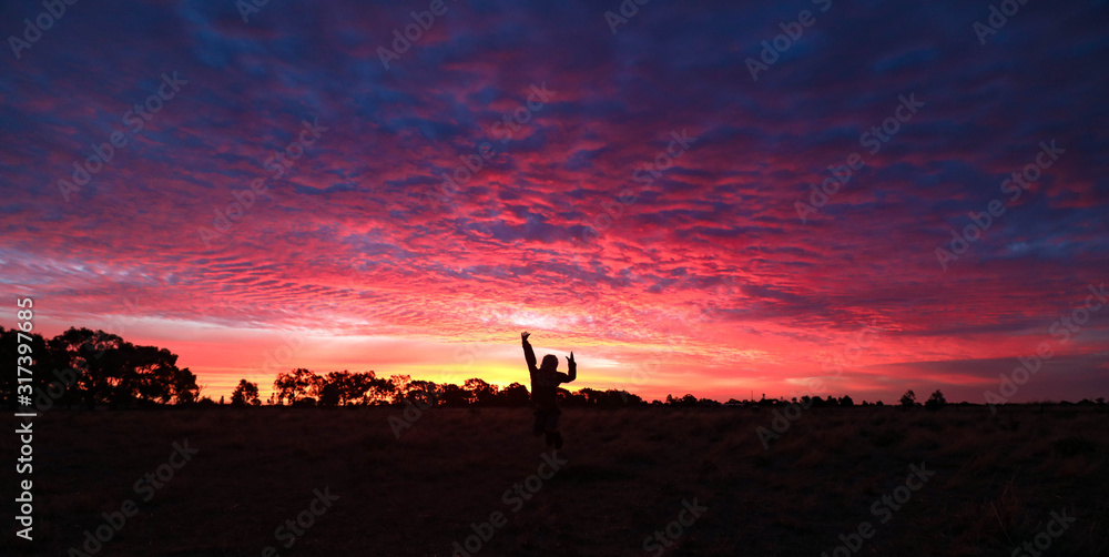 Panoramic image of vibrant pink and purple sunset sky with silhouette of small child jumping in Central Victoria, Australia