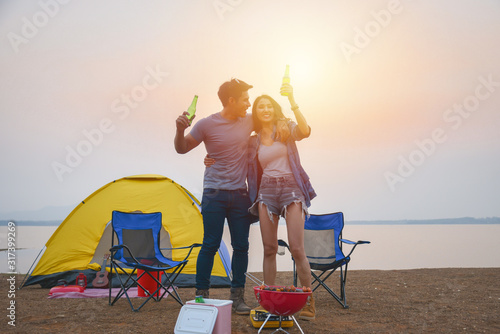 Young asian couple drinking beer and grilling barbecue in front of tent while camping at lakeside