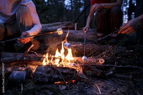 Fototapeta Naklejka Na Ścianę i Meble -  Friends frying sweet marshmallows over a campfire, on a summer evening, in the forest.
