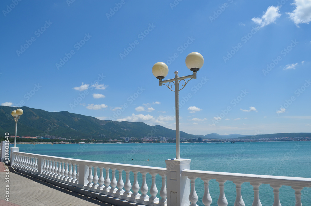 Blue sea embankment with railings and lanterns against the blue sky and hilly mountains.