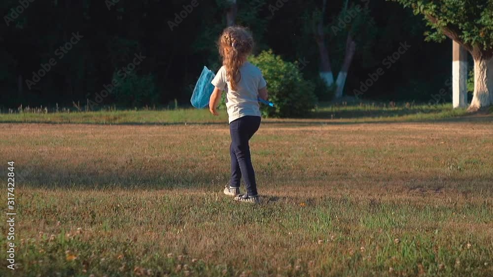 little beautiful blond lady walks holding blue butterfly net in hand on green garden lawn slow motion backside view