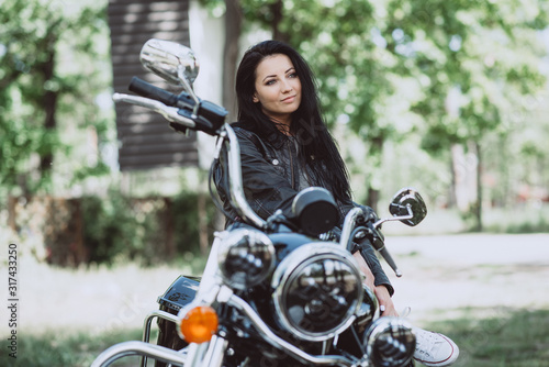 Biker woman in a leather jacket on a motorcycle looks into the distance on a summer sunny day on a green background.