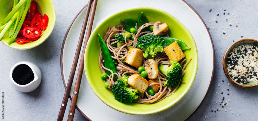 Soba noodles with vegetables and fried tofu in a bowl. Grey background. Top view.