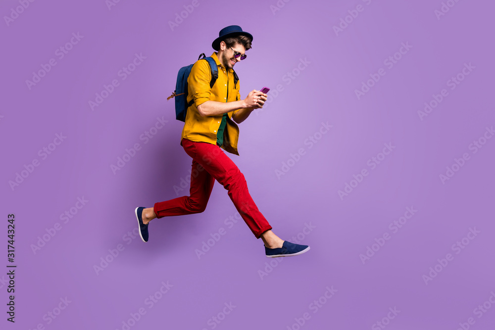 Full size profile side photo of positive hipster guy relax trip jump use smartphone search discounts run fast wear yellow shirt red pants rucksack isolated violet color background