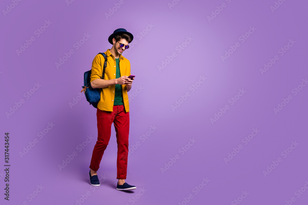 Full length profile side photo of positive guy hipster use smartphone go walk read social network blog news wear yellow shirt red pants bag velvet headwear isolated vibrant color background Full length profile side photo of positive guy hipster use smartphone go walk read social network blog news wear yellow shirt red pants bag velvet headwear isolated vibrant color background