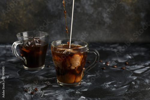   Two glass cups with cold coffee. Black iced coffee in transparent mugs. Streams of coffee and milk pour into a cup and intermix inside. Copy space
