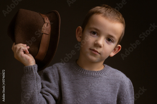 Studio portrait of boy in a gray sweater, on a black background, holding a brown hat in his hand