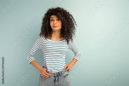 skeptical mixed girl looking at camera while holding hands on hips on grey background