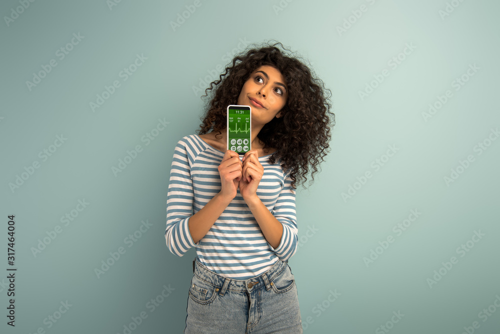 thoughtful mixed race girl looking away while showing smartphone with fitness tracker app on grey background thoughtful mixed race girl looking away while showing smartphone with fitness tracker app on grey background