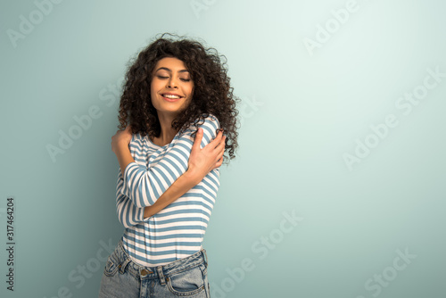 happy mixed race girl hugging herself while standing with closed eyes on grey background