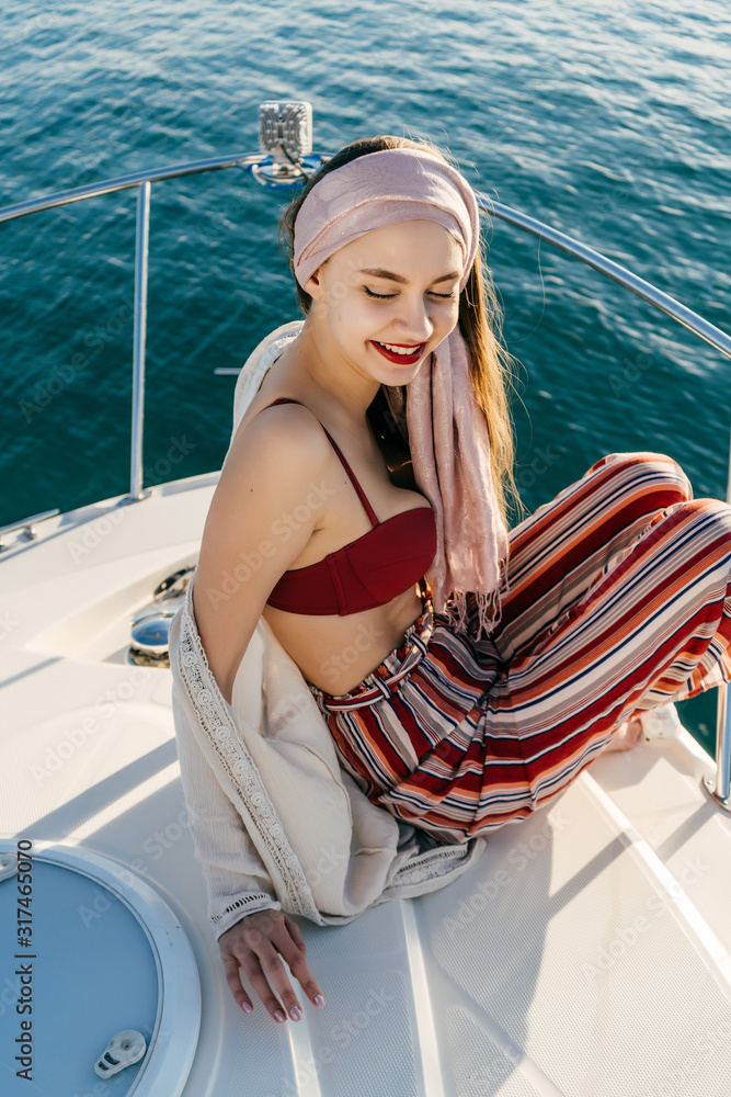 girl in bright striped pants in a bodice from a swimsuit with bright lipstick smiling posing aft of a boat